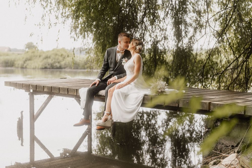 A couple in wedding attire sits on a rustic wooden dock overlooking a tranquil lake with willow trees in the background.