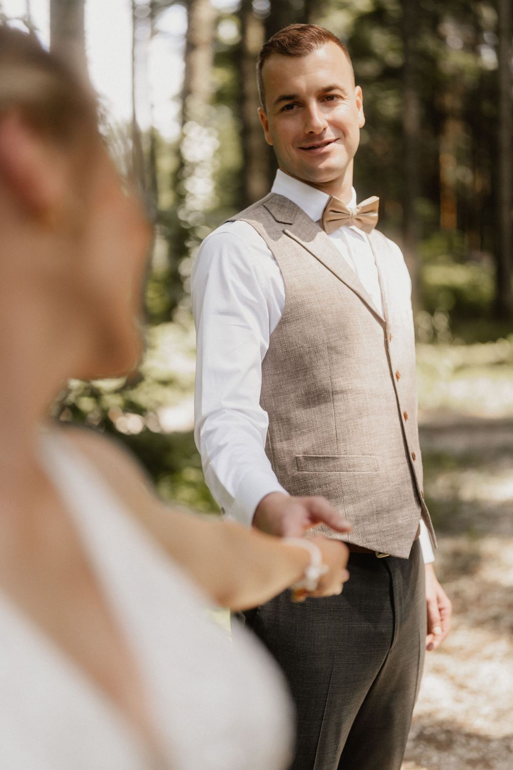 Groom in a beige vest and white dress shirt smiles in an outdoor forest setting.