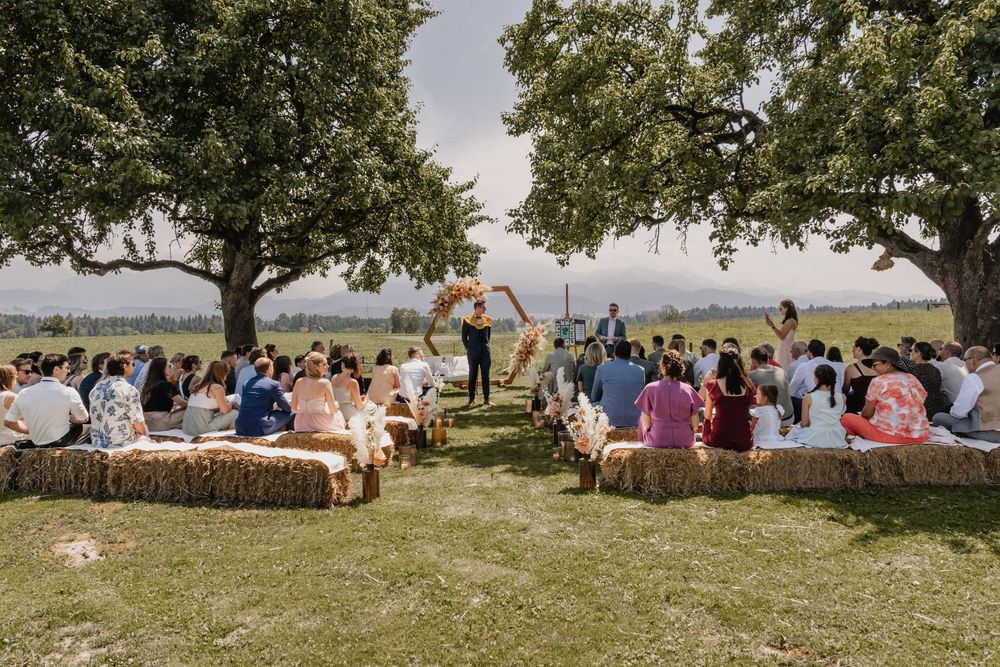 Wedding guests gather under shaded oak trees in a scenic outdoor summer celebration spread across the rolling countryside.