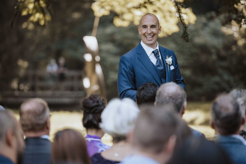 A group gathers outdoors for a wedding ceremony in a garden setting under natural sunlight and greenery.