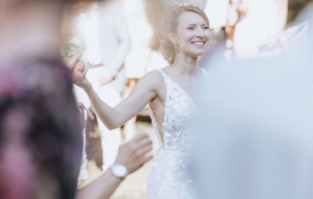 A bride in an elegant white lace dress holds a bouquet while celebrating at an outdoor ceremony.