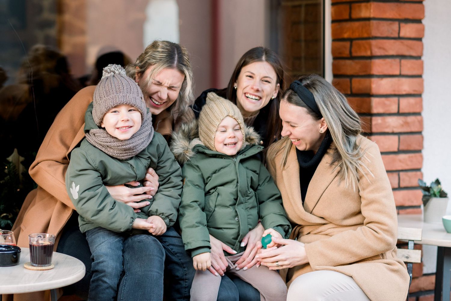 A group enjoying a casual outdoor gathering at a brick-walled cafe wearing winter coats.