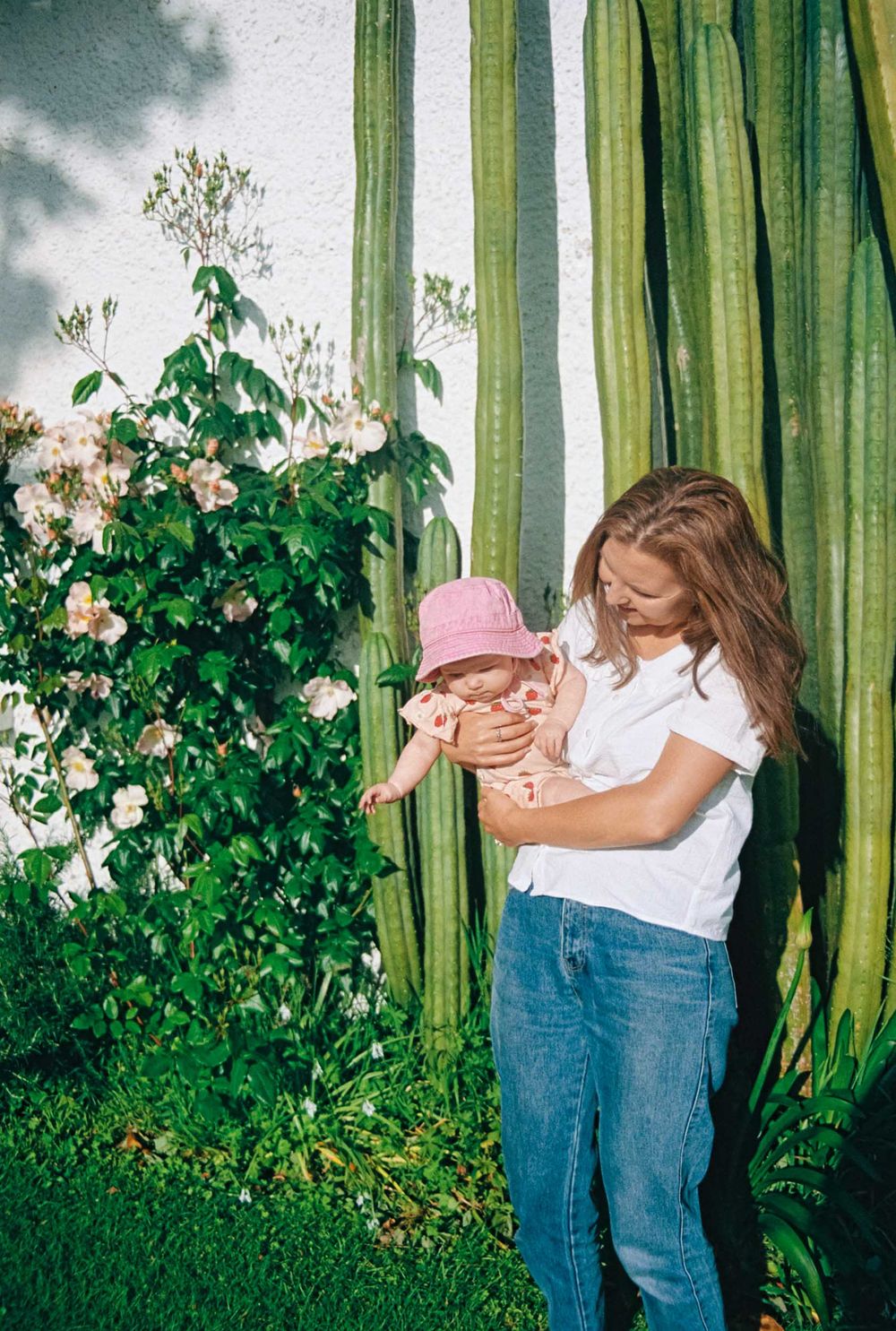 A tender moment captured against green curtains and flowering bushes outdoors with a pink hat under a cloudy sky.