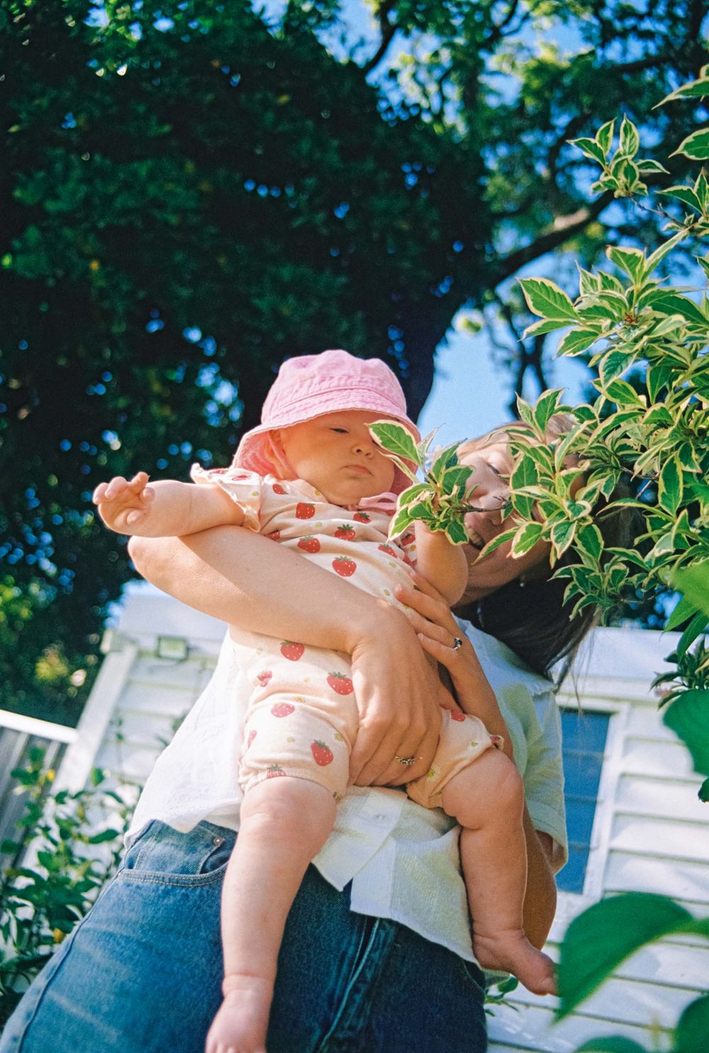 A baby in polka dot dress crawls across someone's lap in a sunny outdoor garden setting with blurred greenery in background.