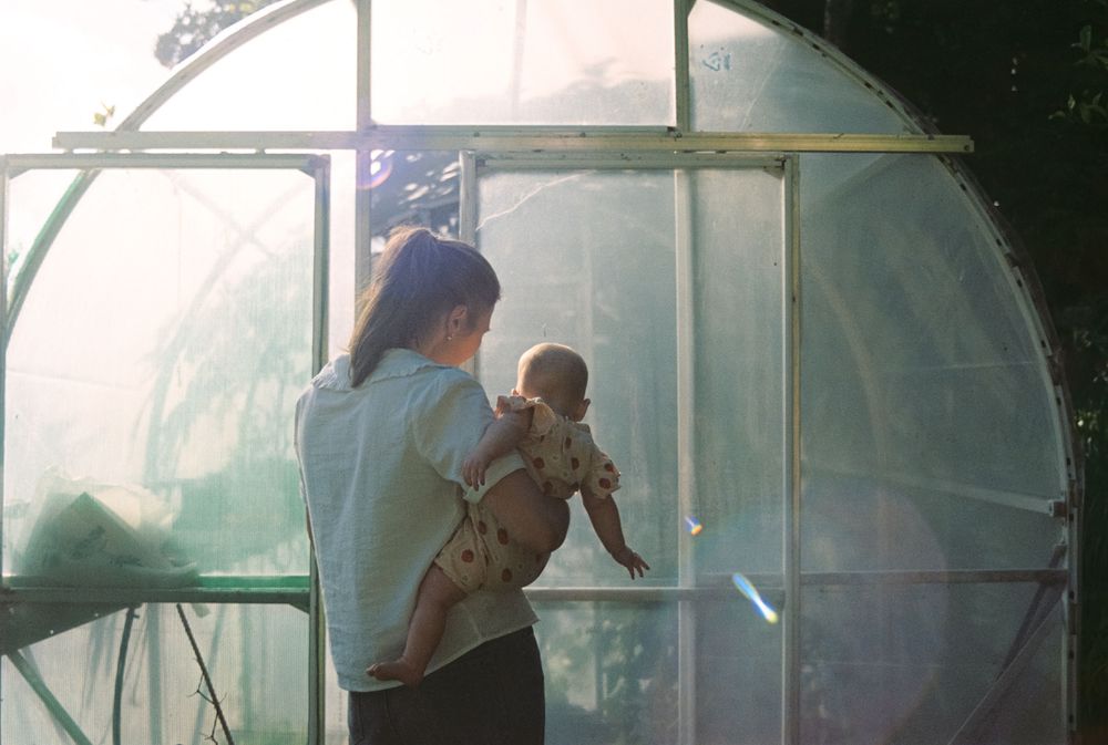 A parent holding a baby while standing inside a greenhouse with sunlight filtering through the translucent walls.