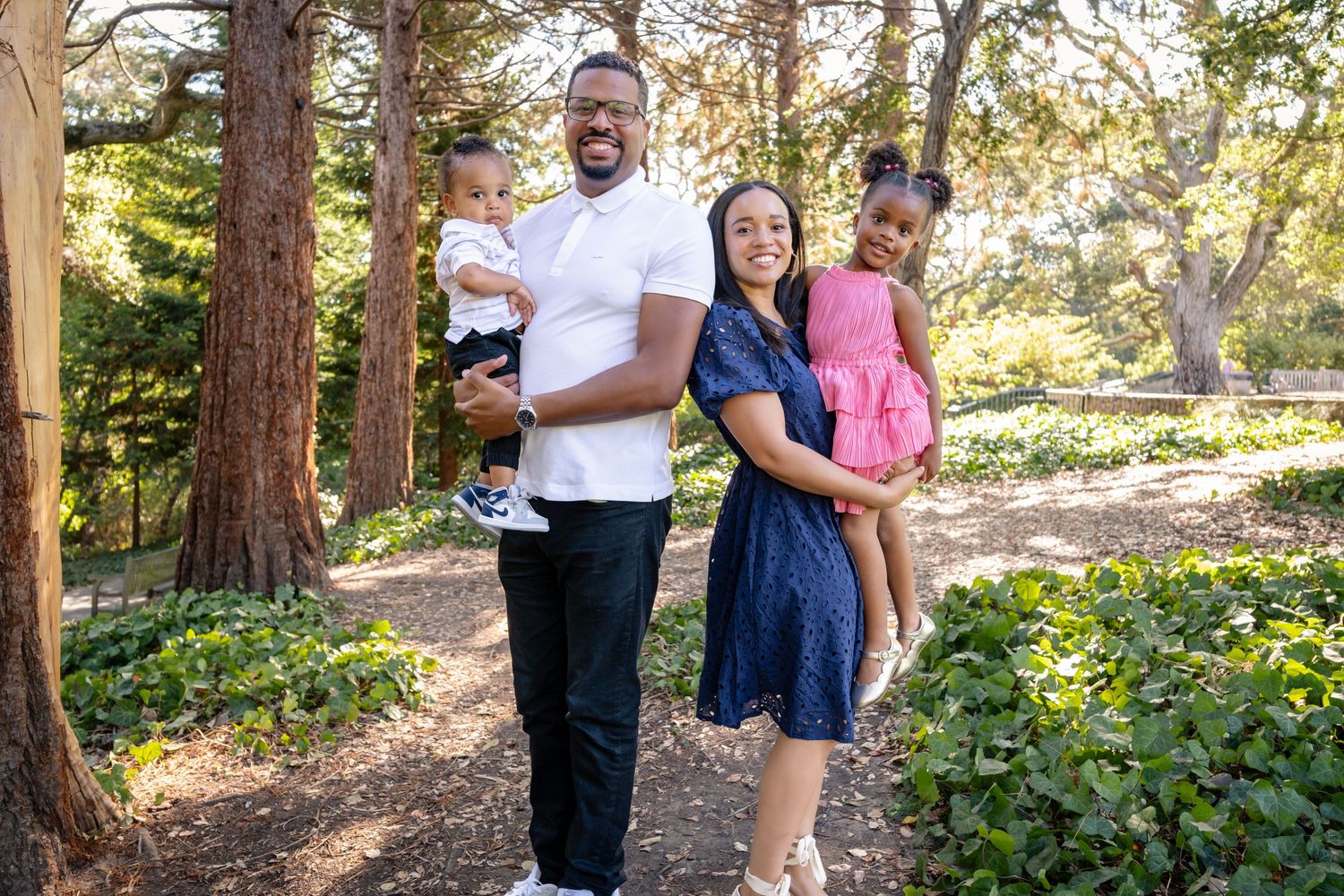 A cheerful family enjoying a sunny day together in a wooded park setting with tall trees in the background.