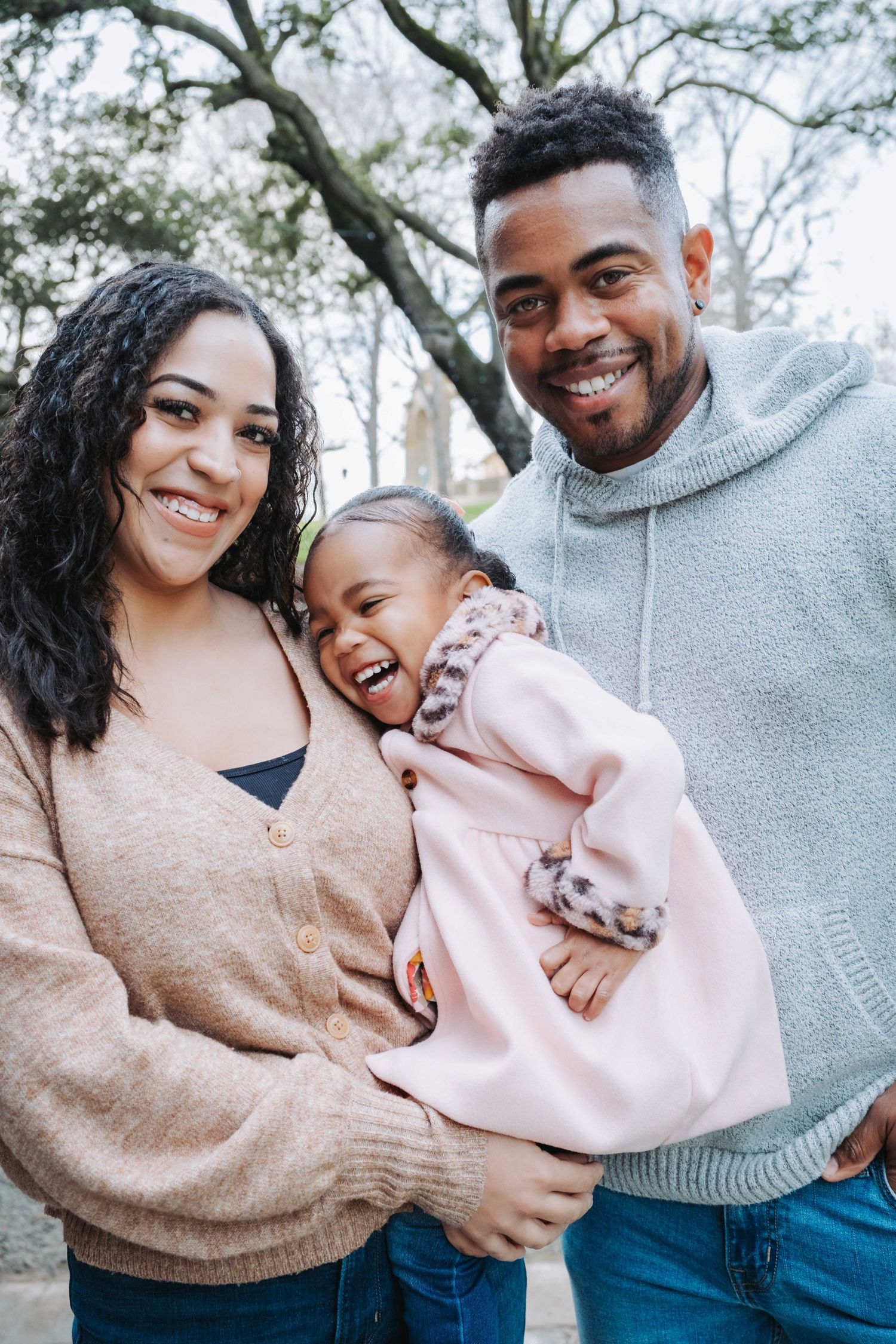 A joyful family of three laughs together outdoors while wearing casual sweaters on a winter day.