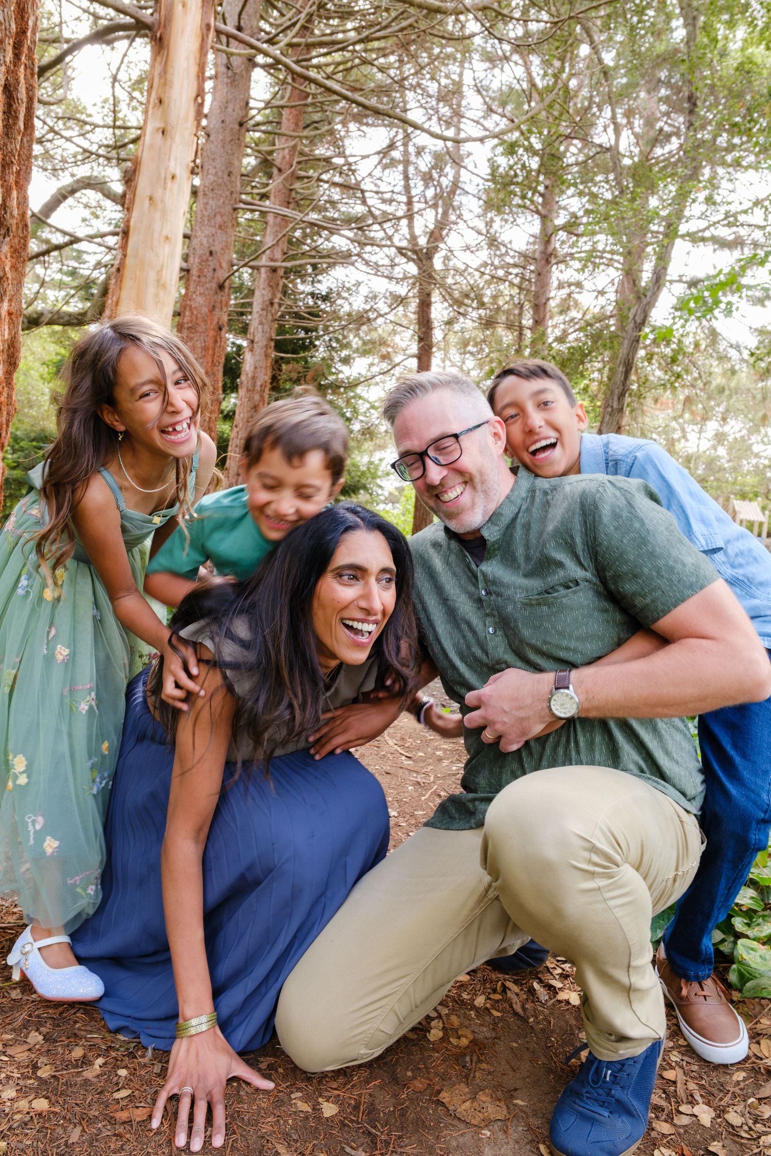 A joyful family of five laughing together outdoors in a forest setting, with parents and three children sharing a fun moment.