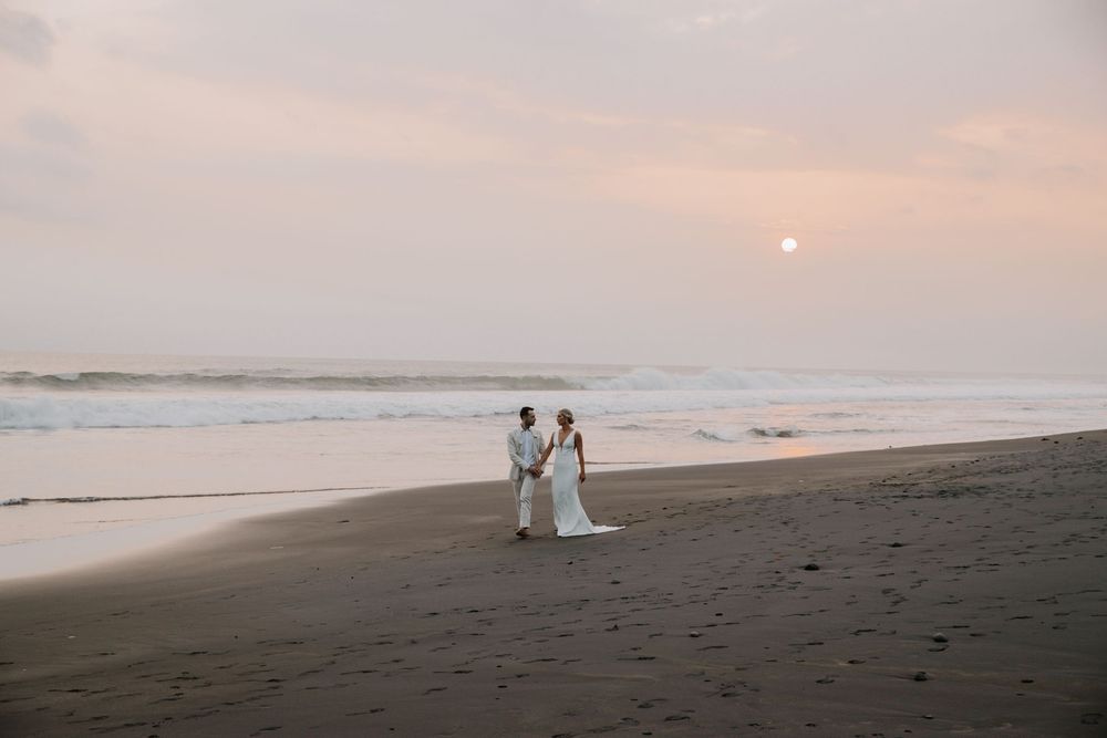A couple in white outfits walks along a Bali beach at sunset as waves crash against the shore.