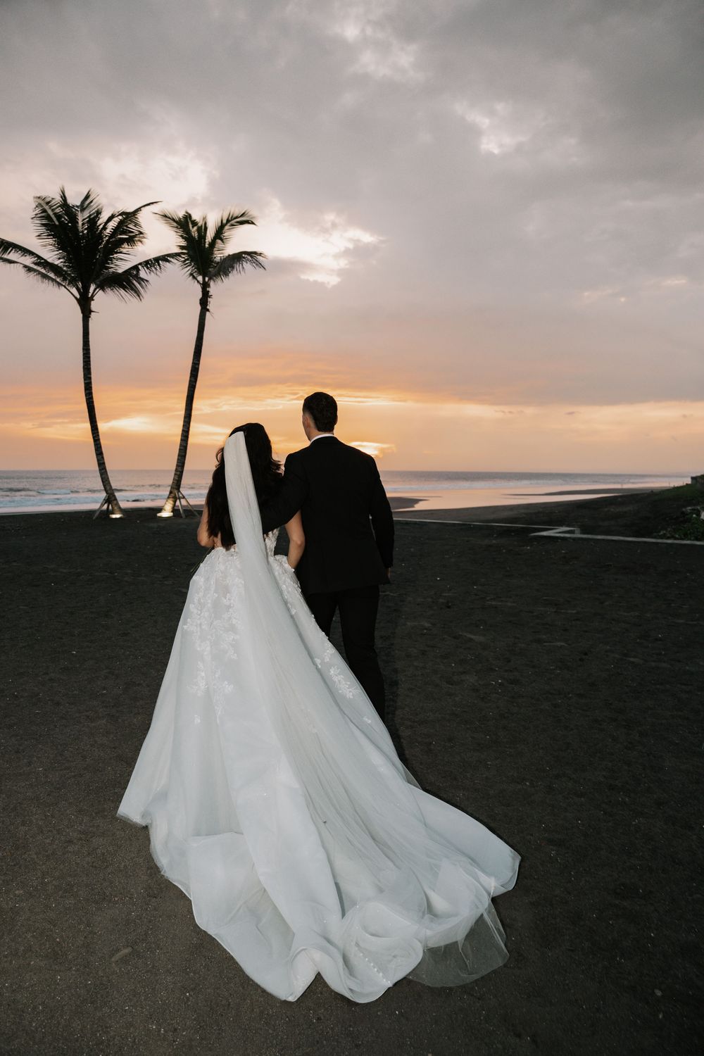 A couple in wedding attire stands on a black sand beach at sunset with palm trees silhouetted against an orange sky.