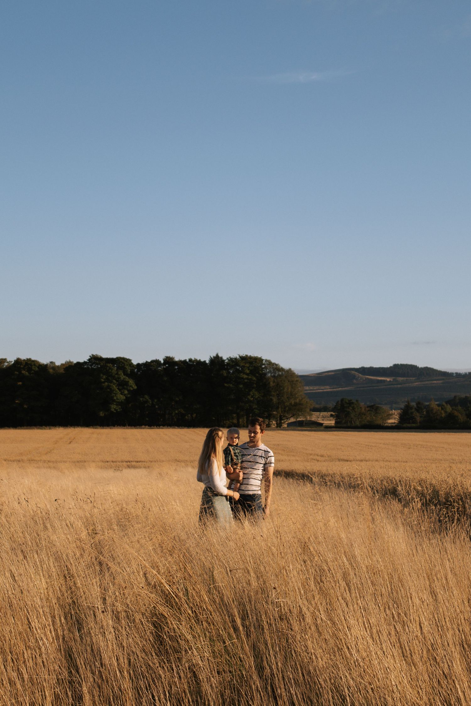 Couple standing together in a golden wheat field with rolling hills in the background.