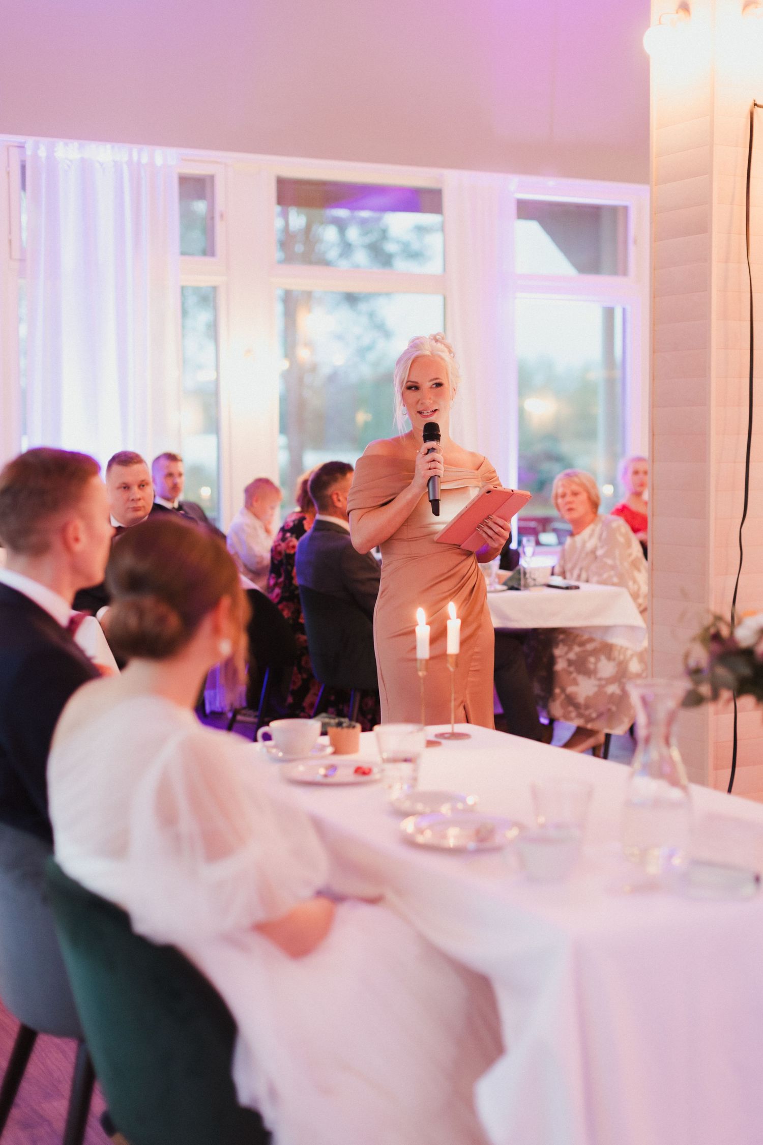 Wedding guests enjoy a lively reception dinner in a bright white room with large windows.