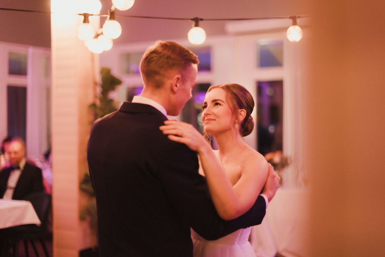 Wedding guests dancing in formal attire under romantic string lights at an indoor reception venue.