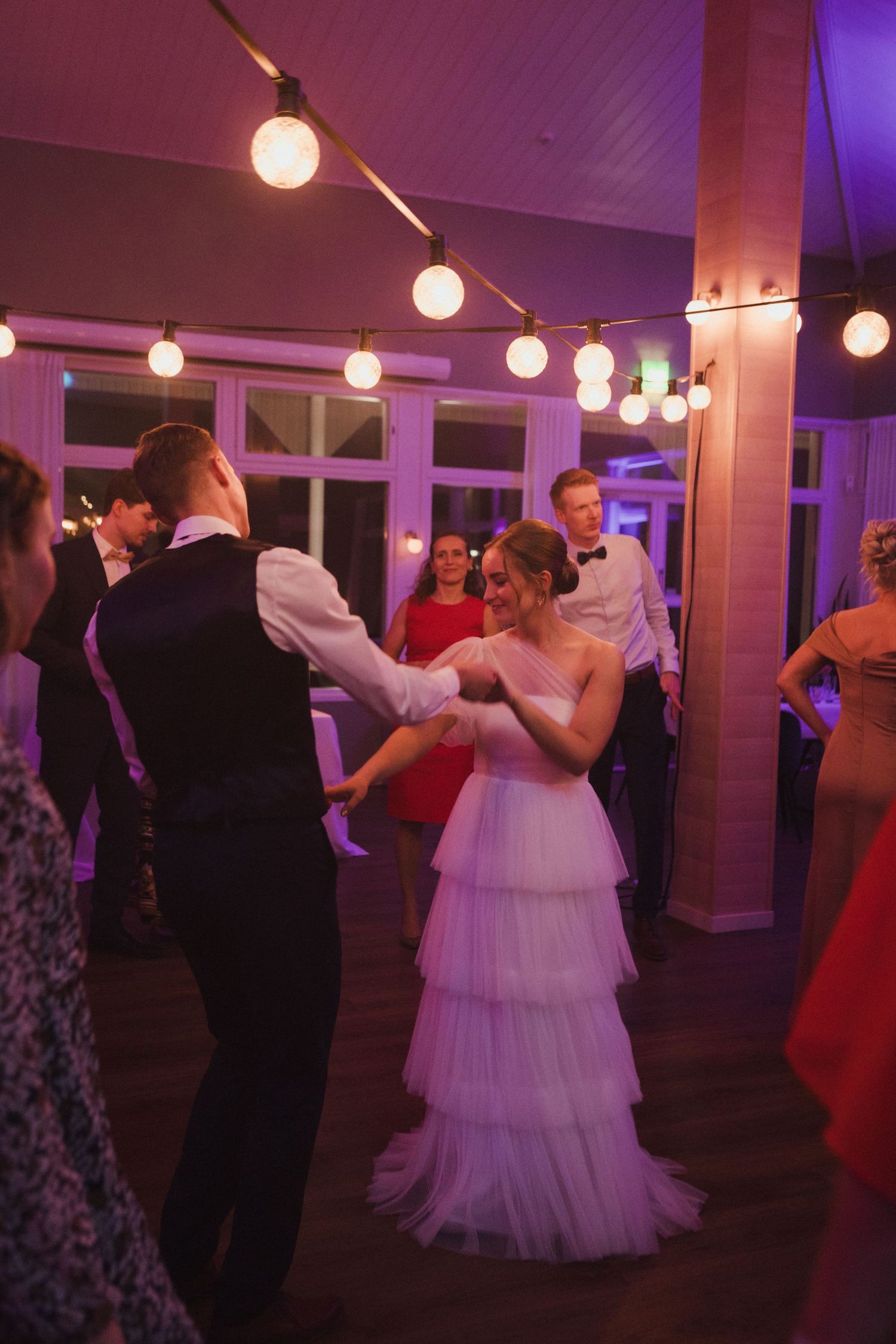 Couples dancing at an evening wedding reception under warm string lights and purple ambient lighting.