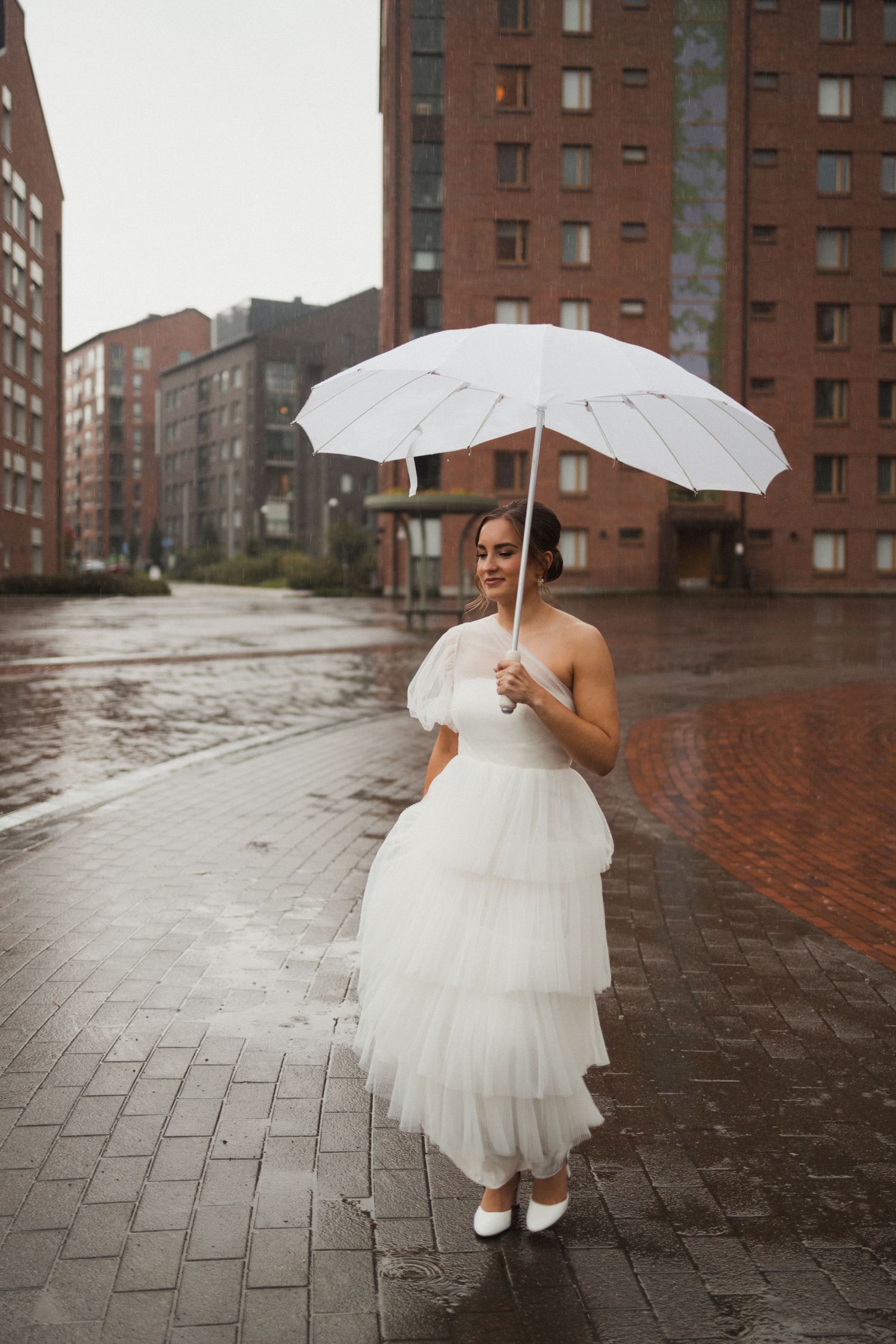 A figure in a white wedding dress holds a white umbrella while walking on wet brick pavement near red buildings.