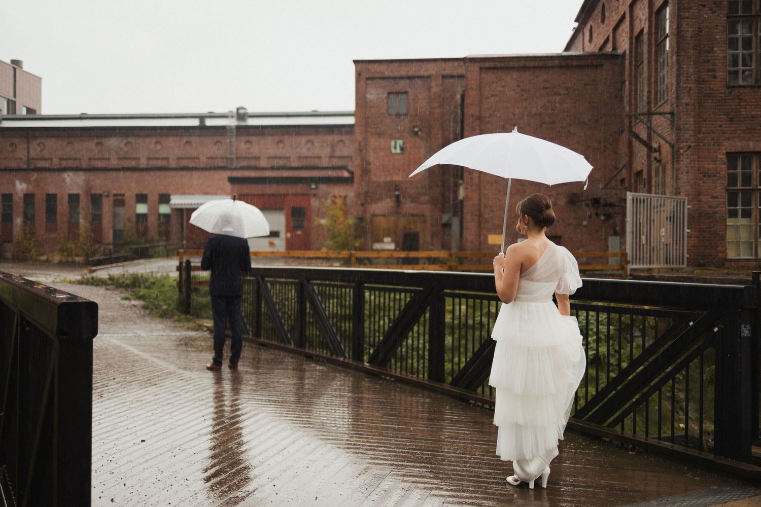 A white-dressed figure with umbrella walks along a rainy canal path beside industrial brick buildings.