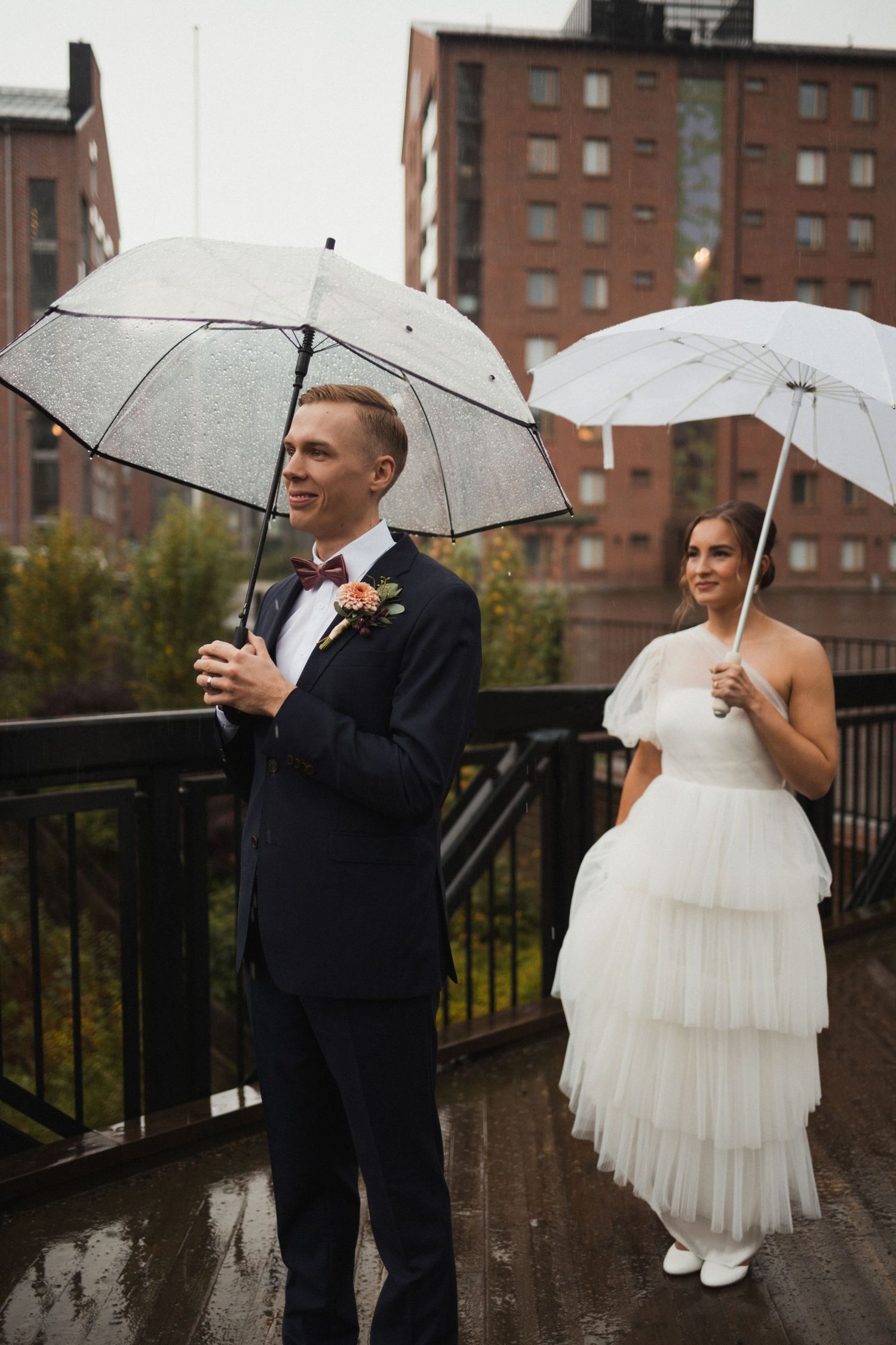Couple sharing romantic moment with white umbrellas on rainy wedding day near brick buildings.