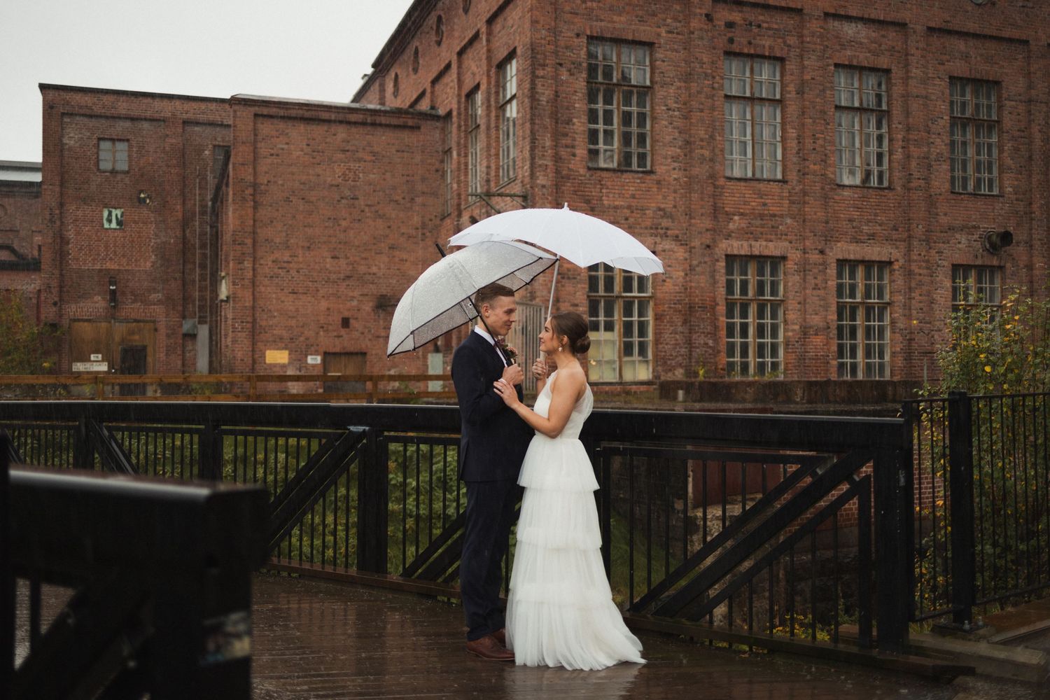 Wedding couple posing in front of a historic red brick industrial building with white umbrellas.