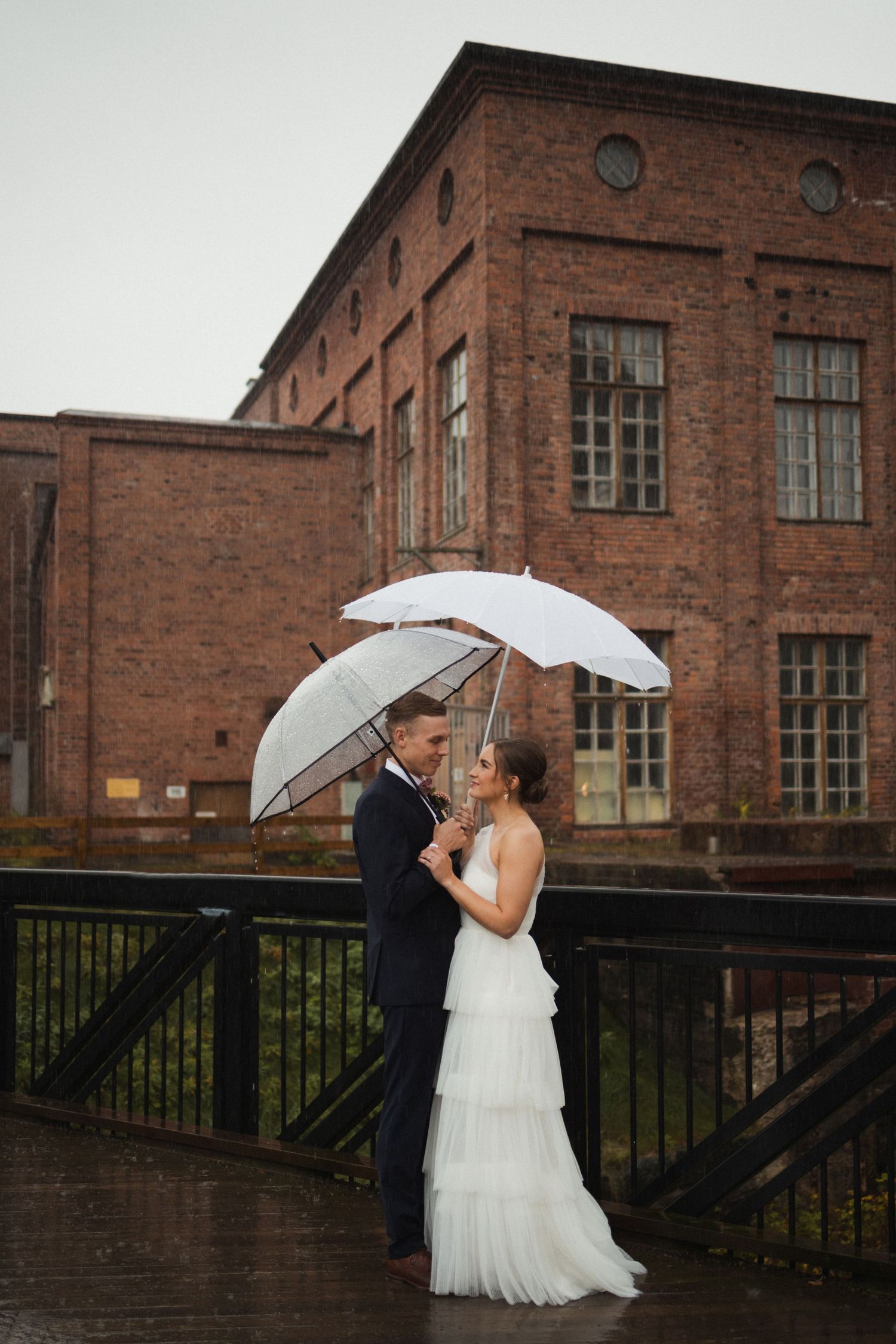 Romantic rainy day wedding photos in front of industrial brick building with white umbrellas.