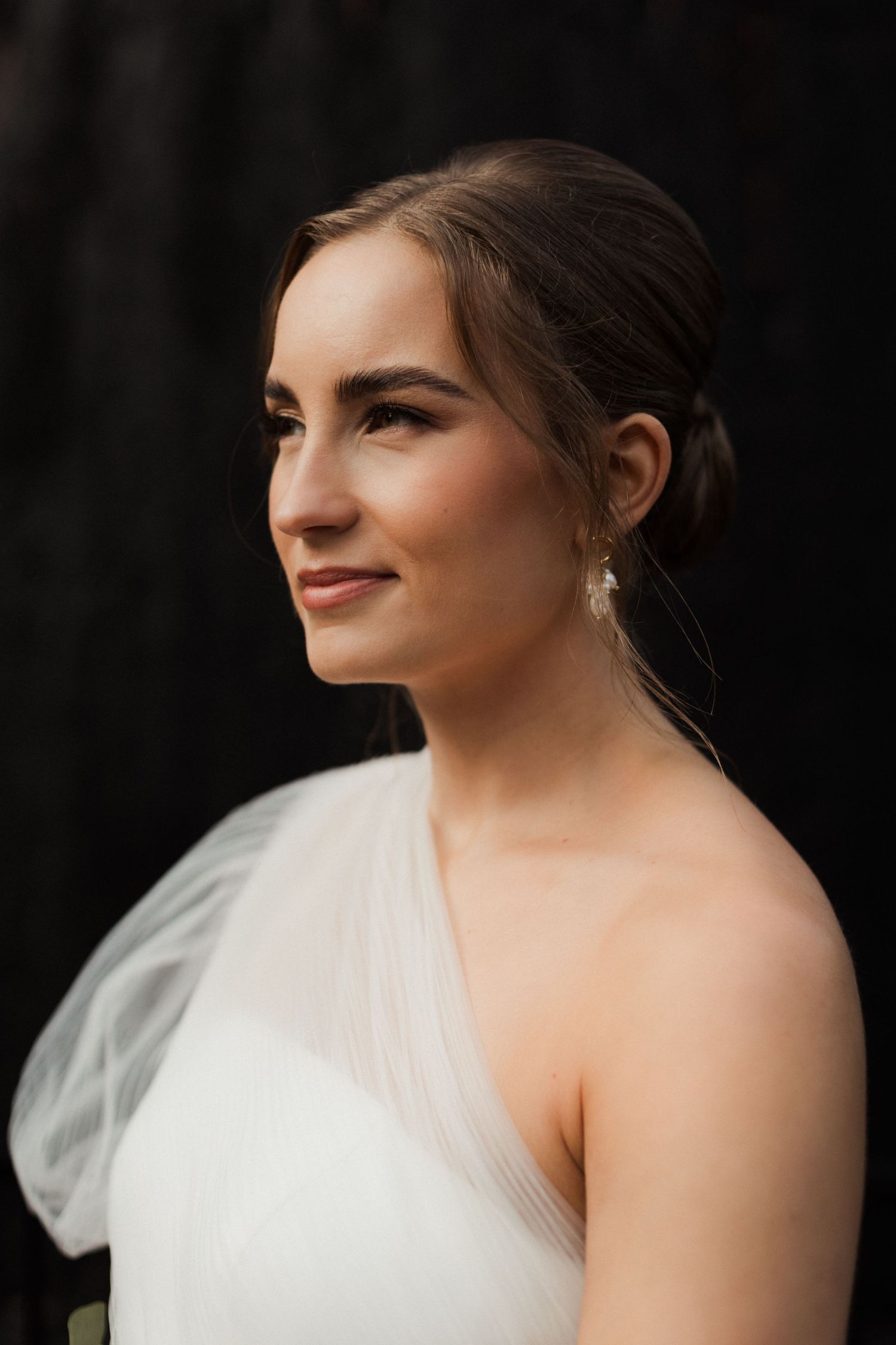 Close-up portrait of a bride in a white wedding dress with delicate tulle details against a dark background.
