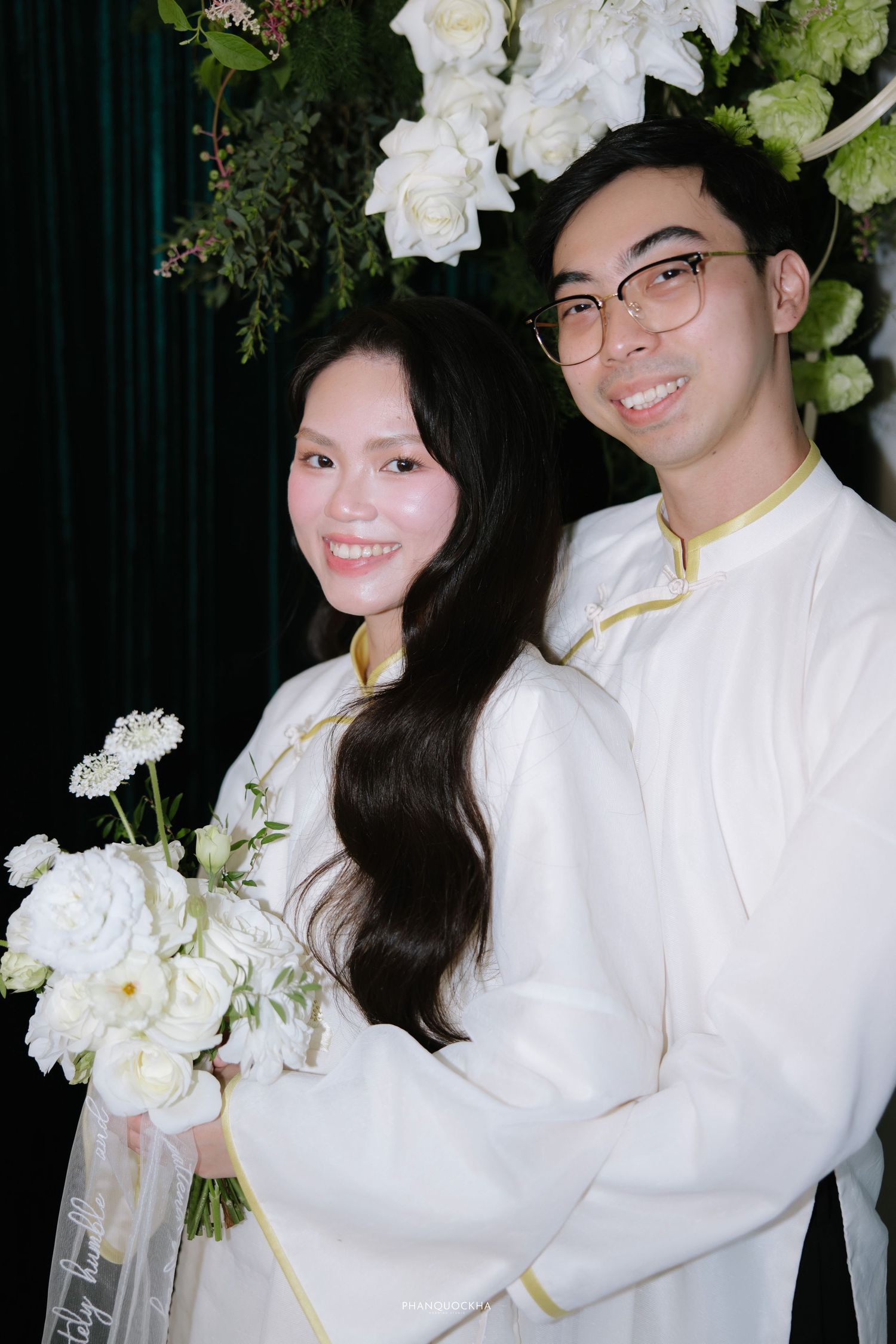 Smiling Vietnamese wedding couple in white ao dai with gold trim pose with white floral bouquet.