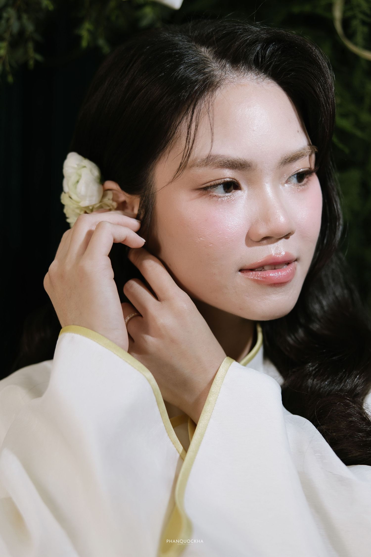 Close-up portrait of a young Asian woman in white ao dai adjusting a white flower in her dark hair.
