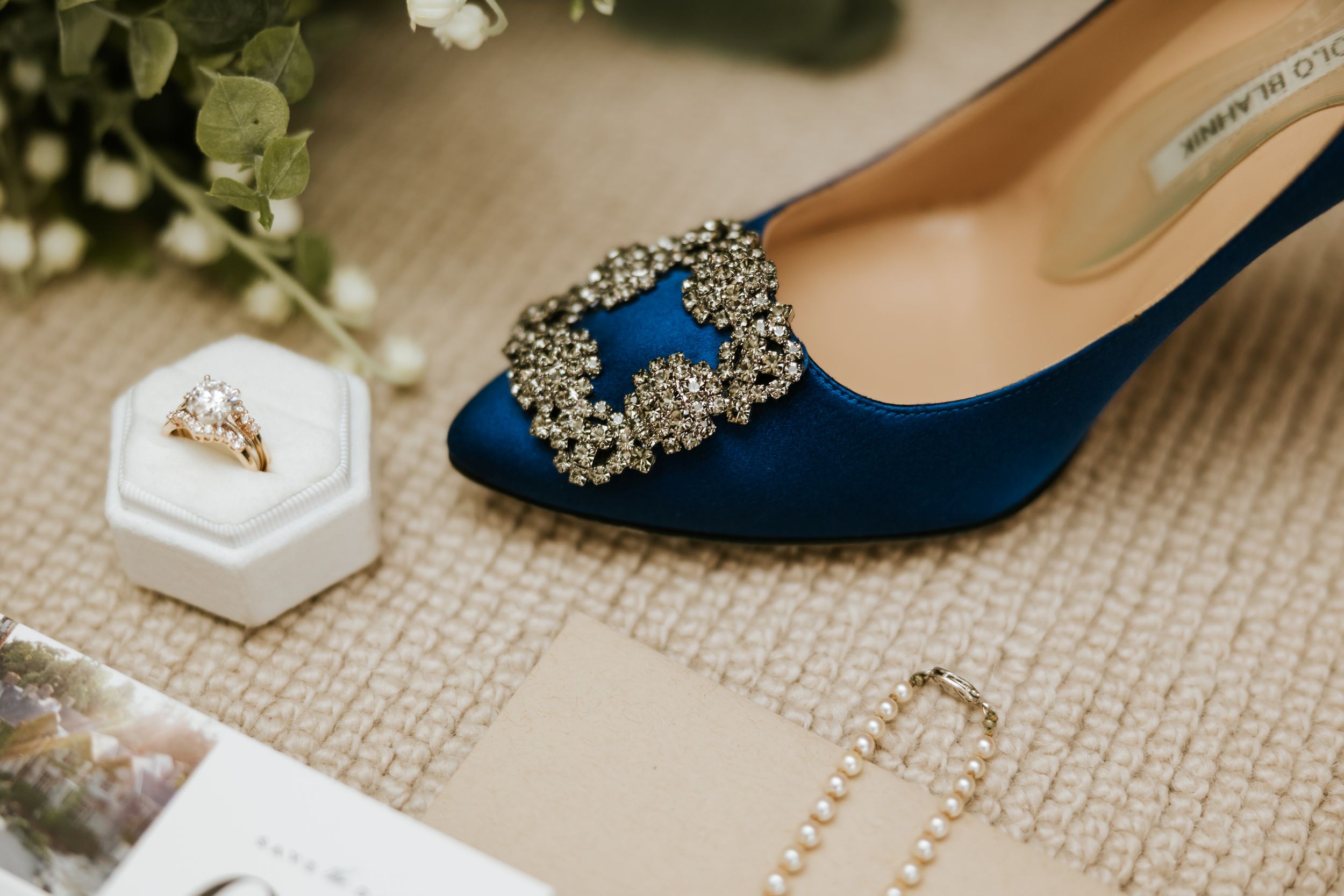 Navy blue bridal shoes with sparkly embellishments sit alongside delicate floral decor and wedding rings on beige carpet.