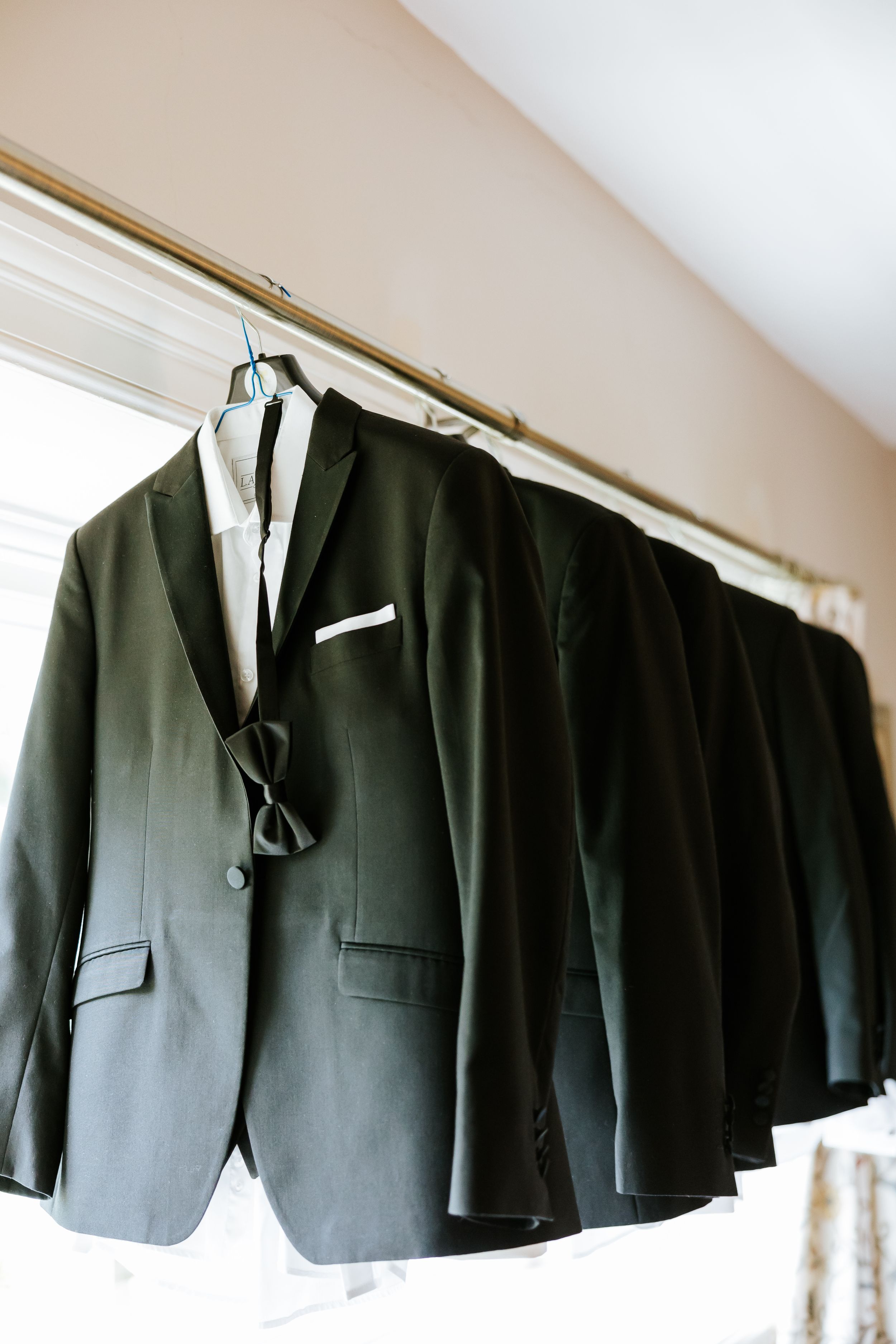Five black formal suit jackets hanging in a row on a rod against a white wall, displaying business attire organization.