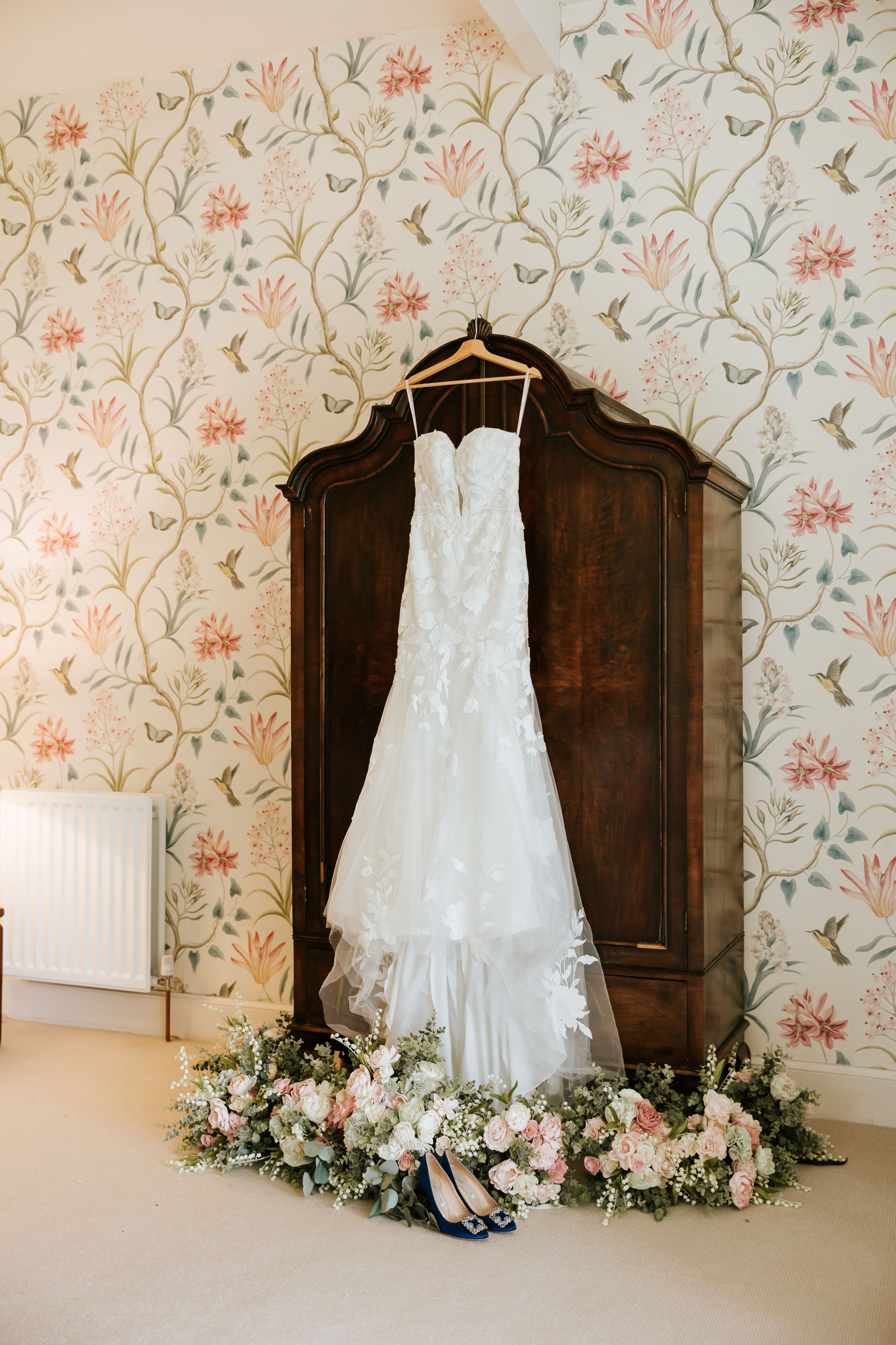White wedding dress hangs against vintage floral wallpaper with dark wooden furniture and delicate floral arrangements.