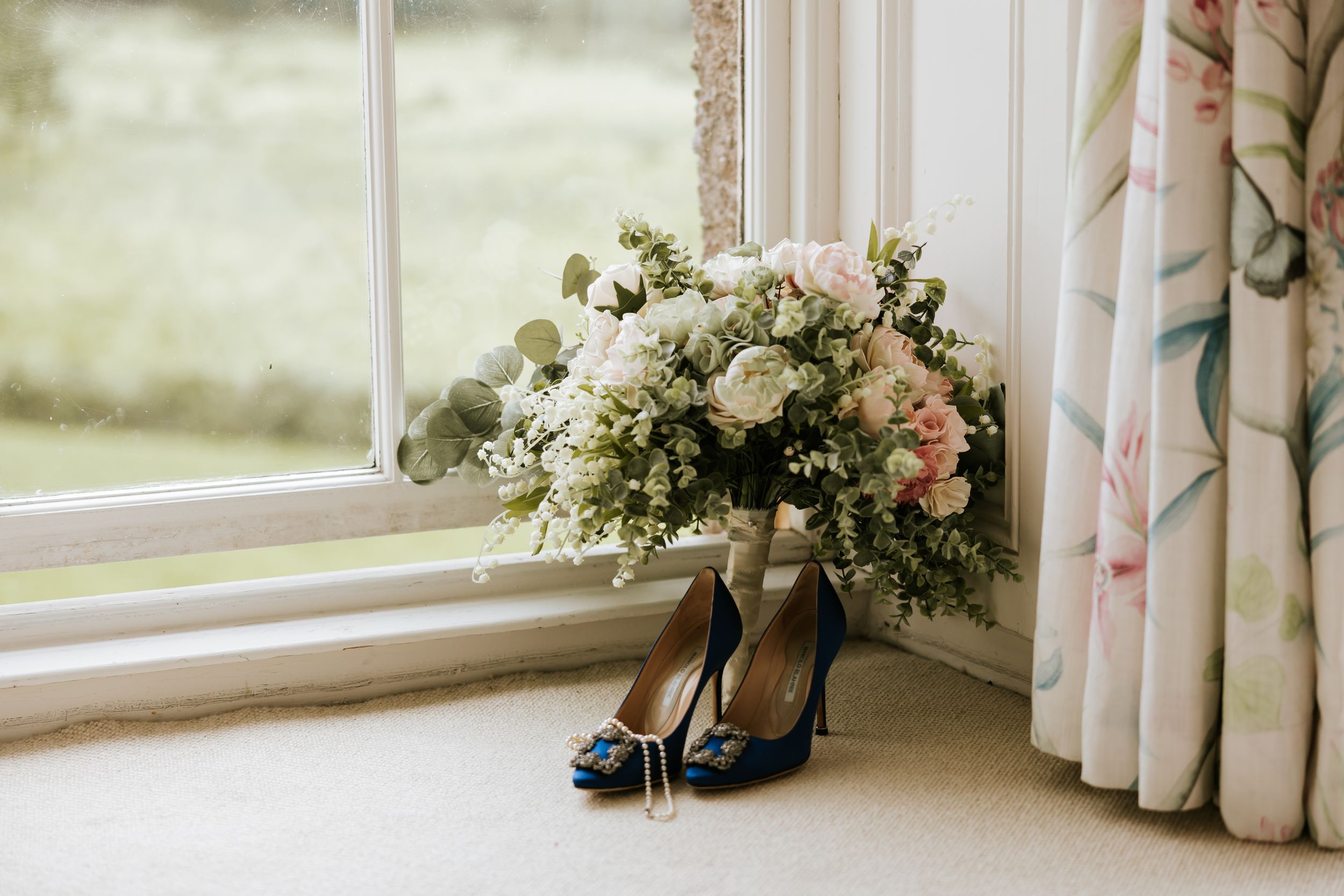 A pair of white bridal shoes sits on a windowsill decorated with delicate floral arrangements and sheer curtains.