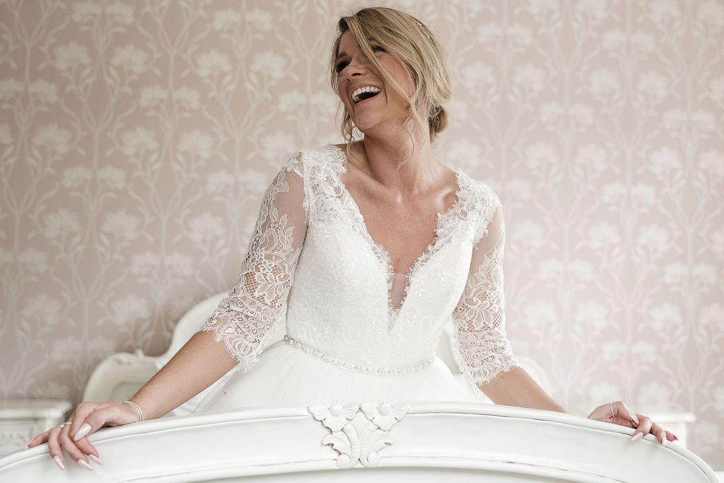 Joyful bride laughing while sitting in the bright Bridal Suite at Kelham House wearing a white lace long-sleeved wedding dress