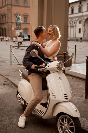 A couple sharing romantic moments on a white Vespa scooter in a European plaza.