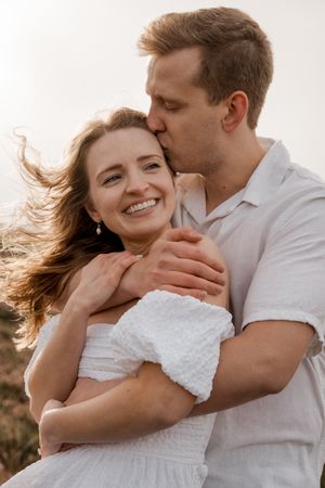 A romantic couple embraces outdoors in white clothing as the wind gently blows through her hair.