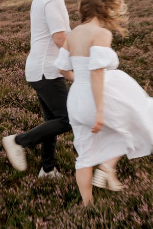 A couple in wedding attire dances playfully in a field of purple wildflowers at sunset.