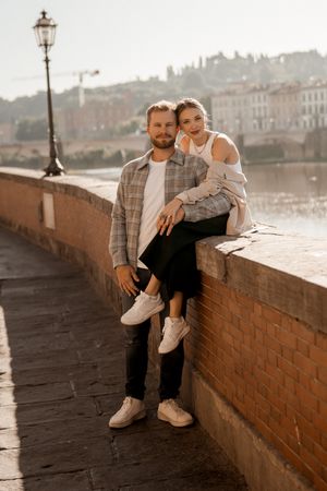 Couple posing casually by riverside in Florence with historic architecture in background.