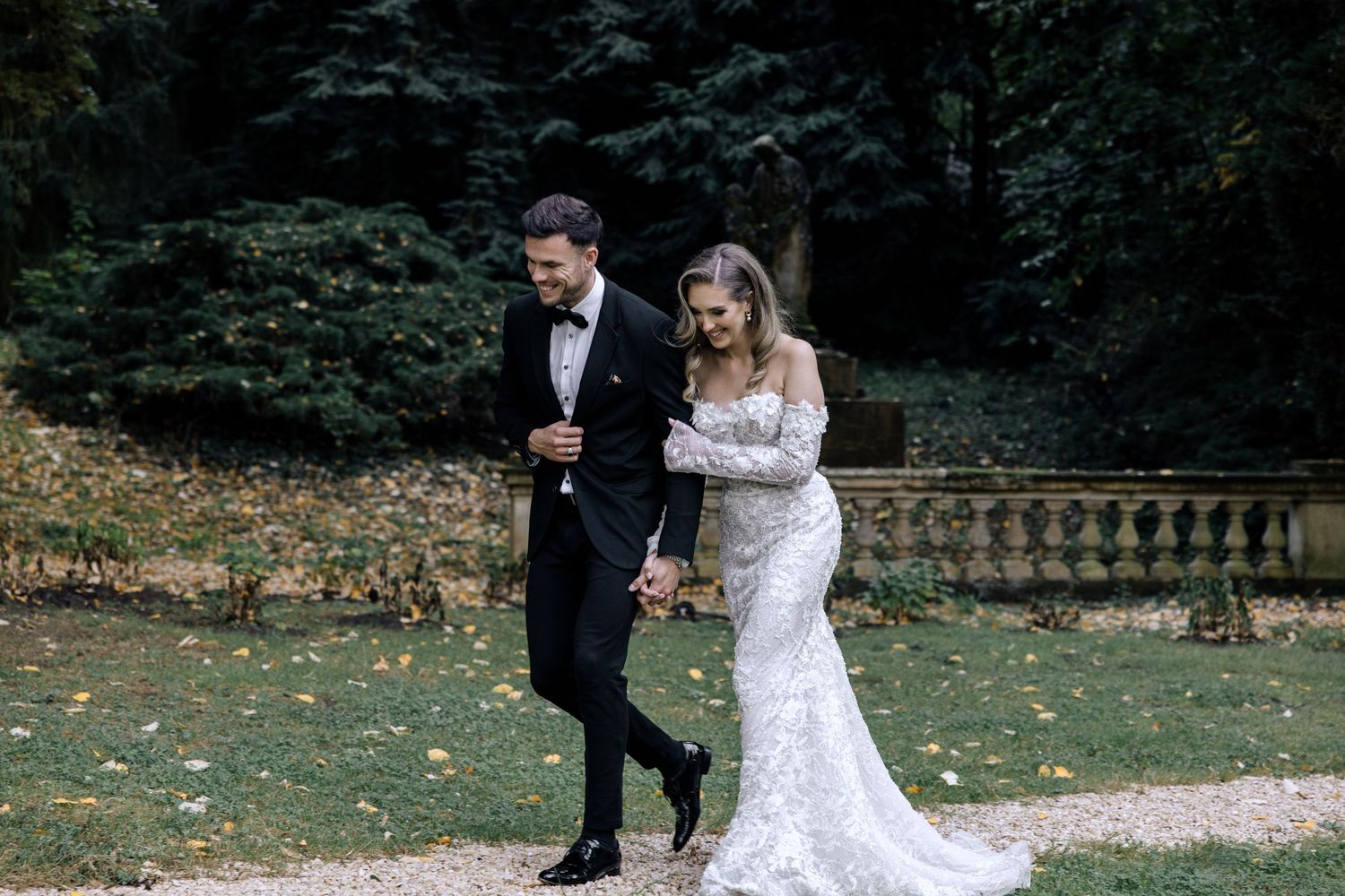 A couple in wedding attire walks along a scenic garden path surrounded by lush greenery and autumn leaves.