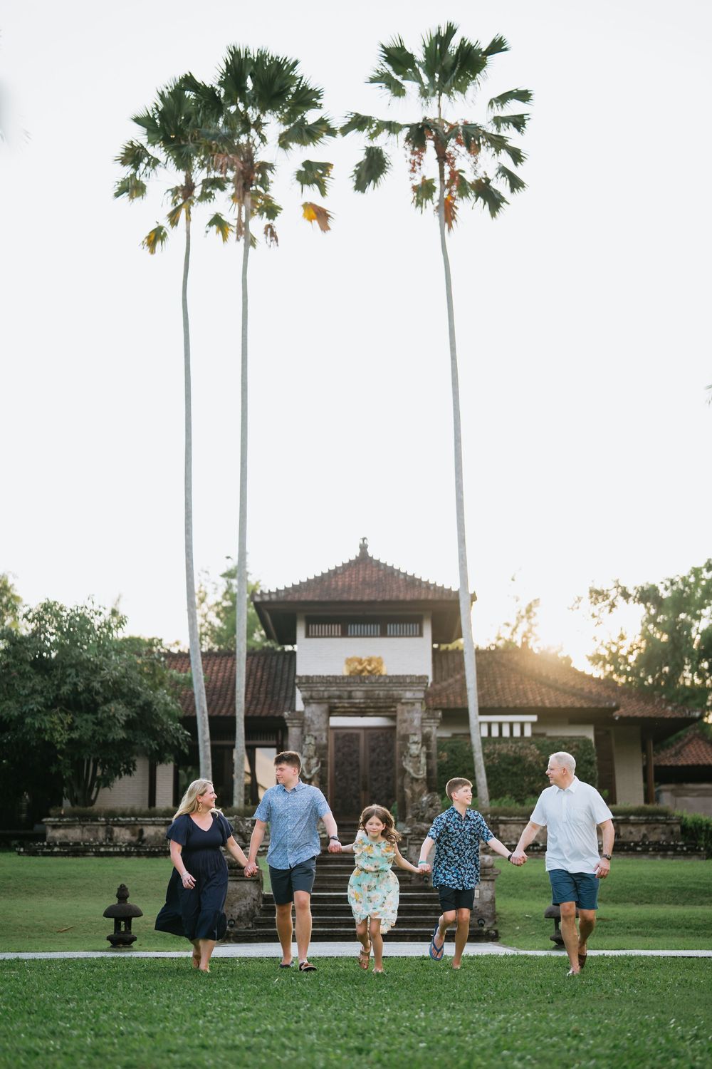 A group walks together on a grassy lawn with tall palm trees and a pagoda-style building in the background at sunset.