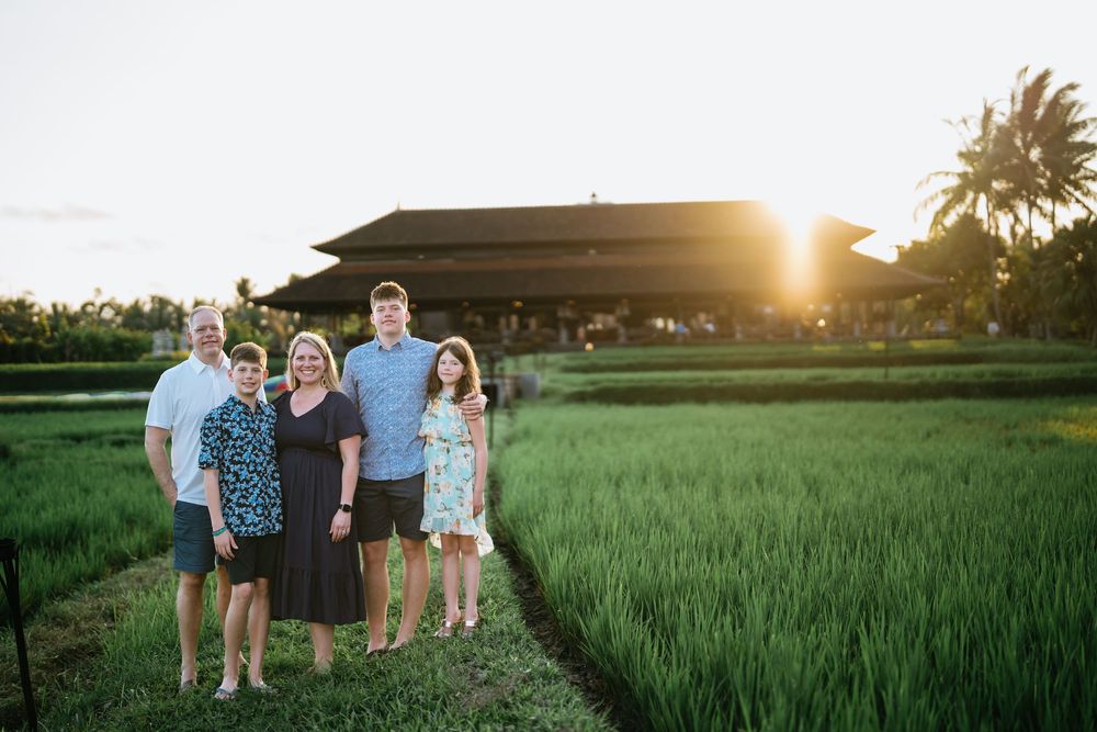 A family poses together on a path through rice fields with a traditional building and sunset in background.