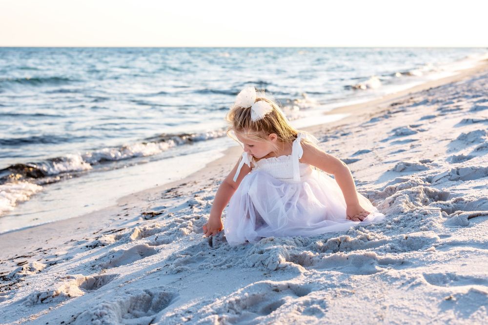 A person in a white dress sits on a sandy beach near the ocean during golden hour.