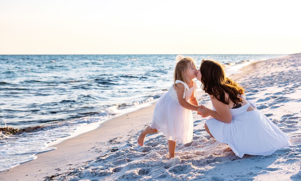 Two people in white dresses walk together along a sandy beach at sunset with ocean waves in the background.