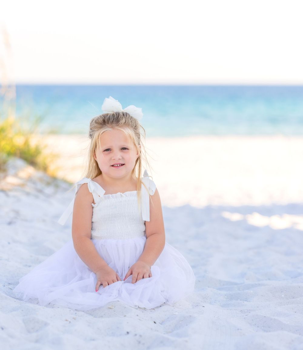 A small child in a white dress plays in soft white sand on a sunny beach with blue sky and water in background.