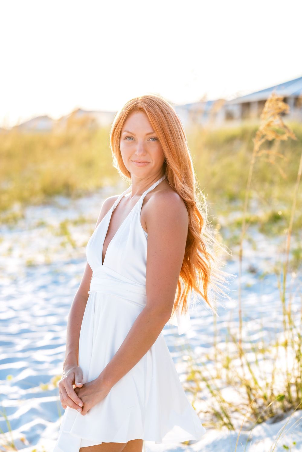 A person in a flowing white dress poses on a sandy beach during golden hour sunset while sea oats sway in the breeze.