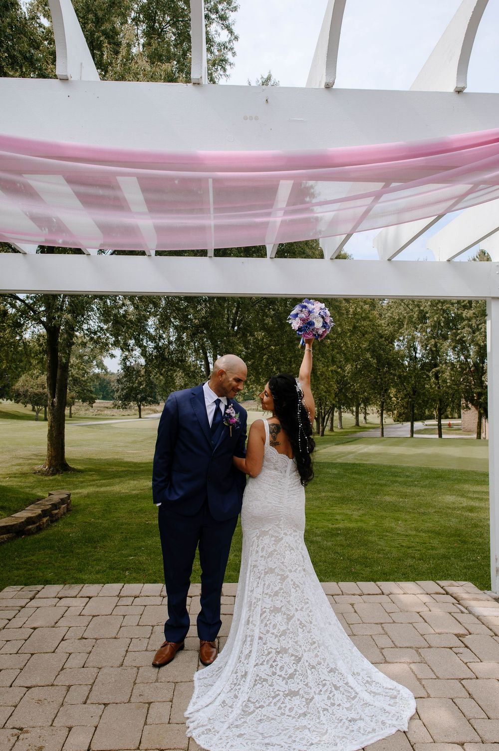 A couple celebrates their wedding day under a white pergola with pink draping at an outdoor venue on a sunny afternoon.