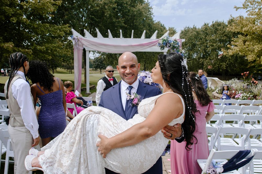 Wedding guests gather around a white wooden gazebo and chairs during an outdoor ceremony at a park venue.