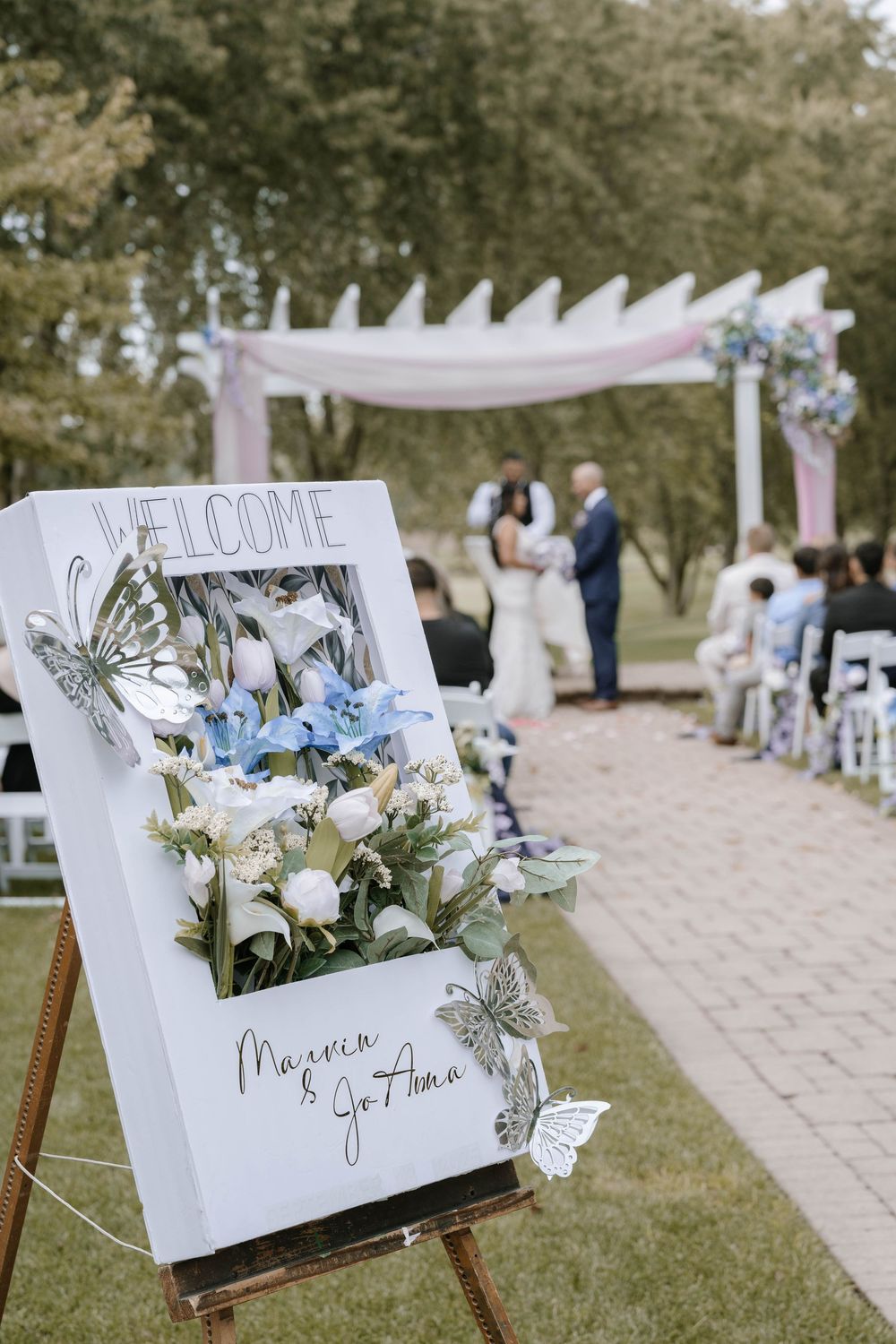 Welcome signage for wedding ceremony displayed on easels with white and blue floral arrangements.