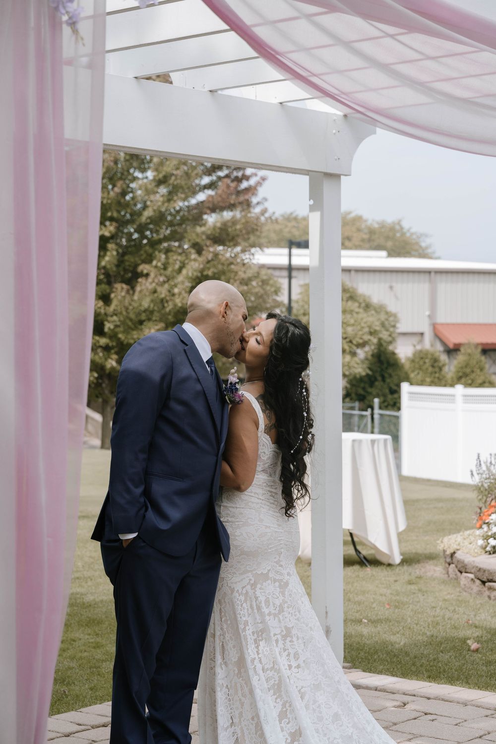 Couple sharing a romantic kiss under an elegant white pergola with pink drapery during their wedding ceremony.
