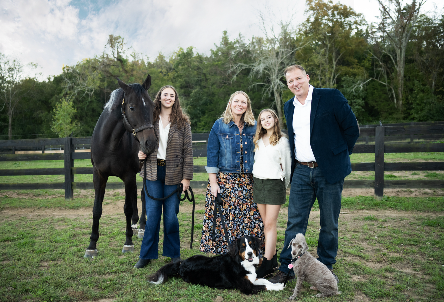 Group standing in pasture with a black horse and small dog at wooden fence during outdoor photo session.
