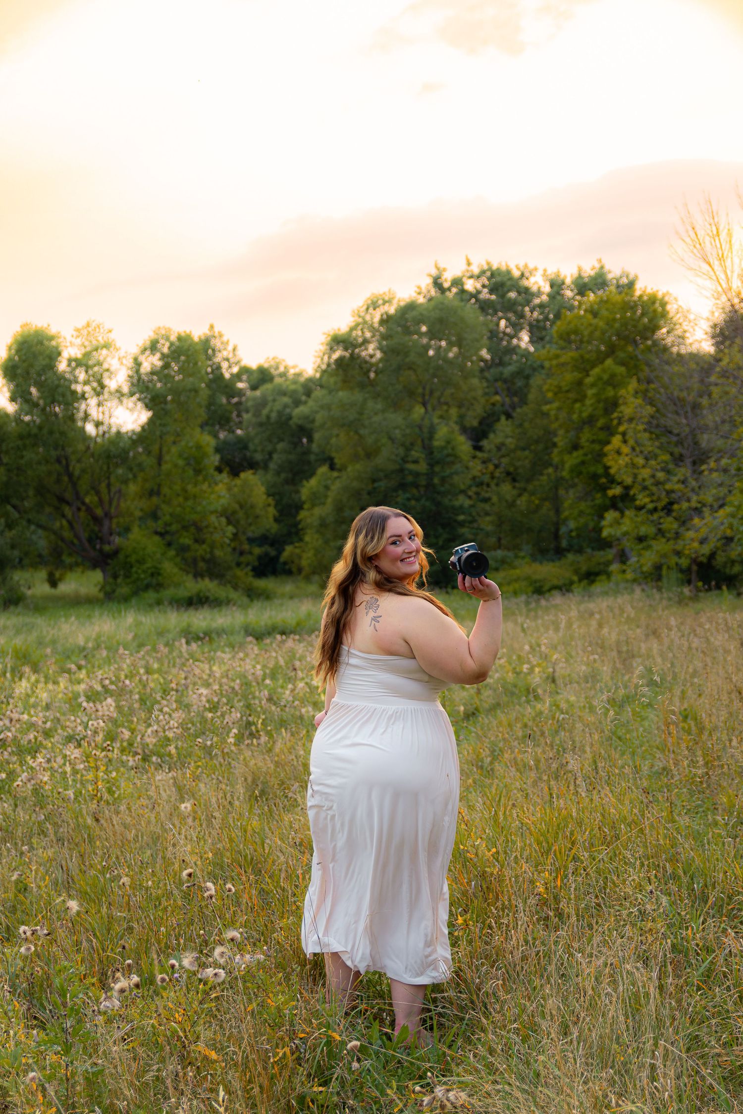 Young woman in cream bow sweater and pink skirt smiling outdoors at golden hour near a tree.