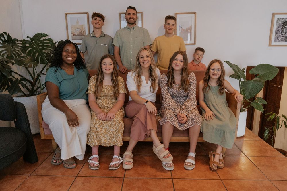 A group of friends sits together wearing coordinated sage green and floral print outfits in a cozy home setting.