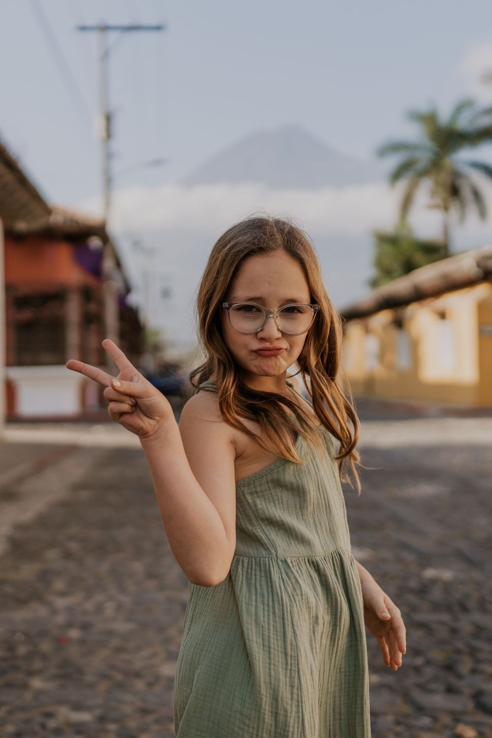 Person in a green sundress making peace sign on cobblestone street with palm trees and mountain in background.